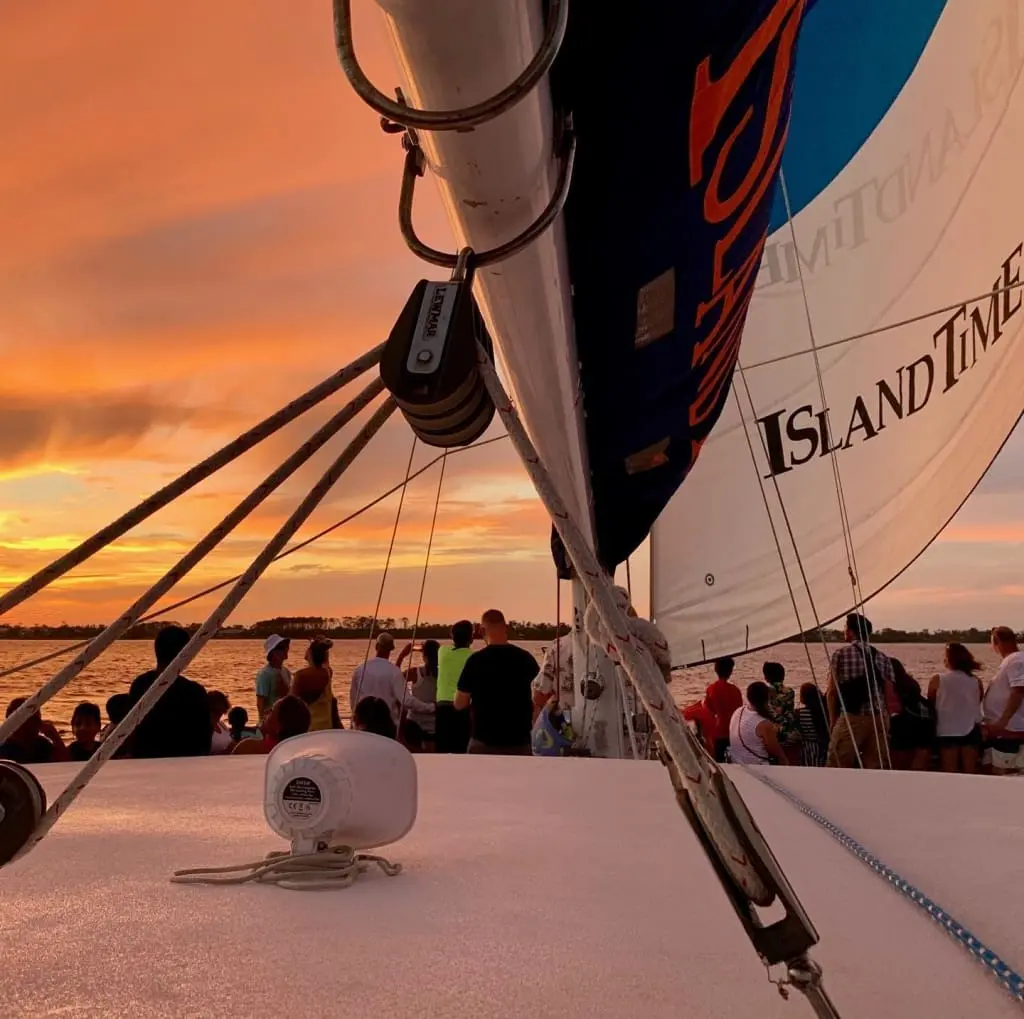 Passengers gathered on a sailboat deck watching a stunning sunset during a dolphin cruise in Panama City, FL, with vibrant orange sky and 'Island Time' sail visible.