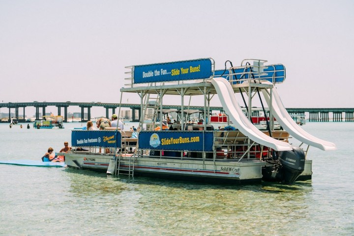 Boat with two slides on water near a bridge, people on deck and a floating mat.
