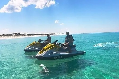 Two people riding jet skis on clear blue ocean near a sandy beach.