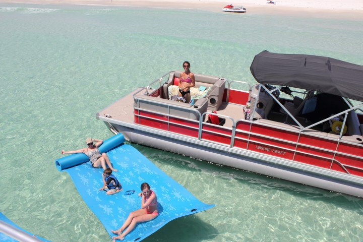 People relax on a pontoon boat with two lounging on a floating mat on clear water.