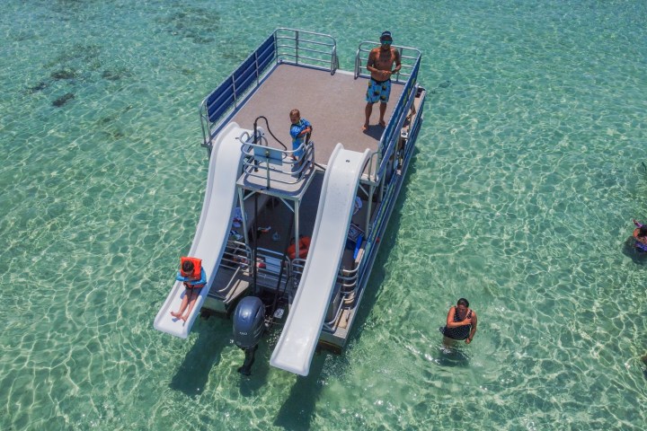 Pontoon boat with slides on clear water, people swimming nearby.
