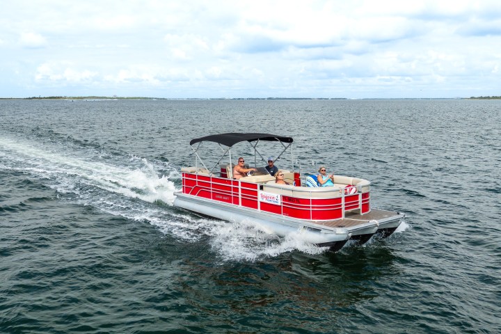 Red pontoon boat with people cruising on open water under cloudy sky.
