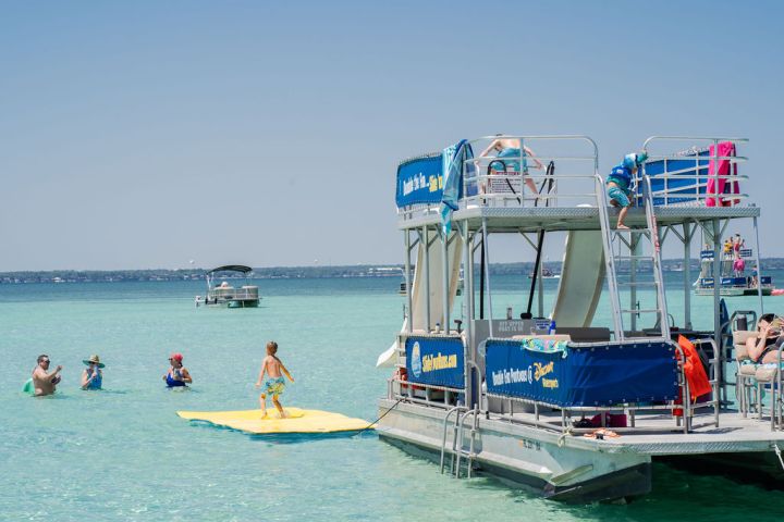 Children playing on a floating mat near a double-deck boat in clear water.