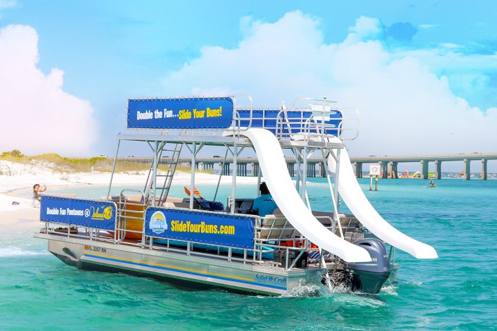 Double-deck boat with slides on turquoise water near a bridge.