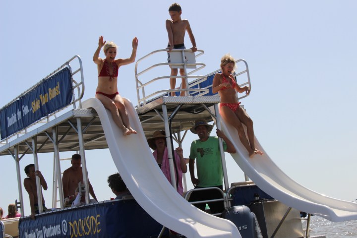 Children sliding down two water slides on a boat with adults supervising.