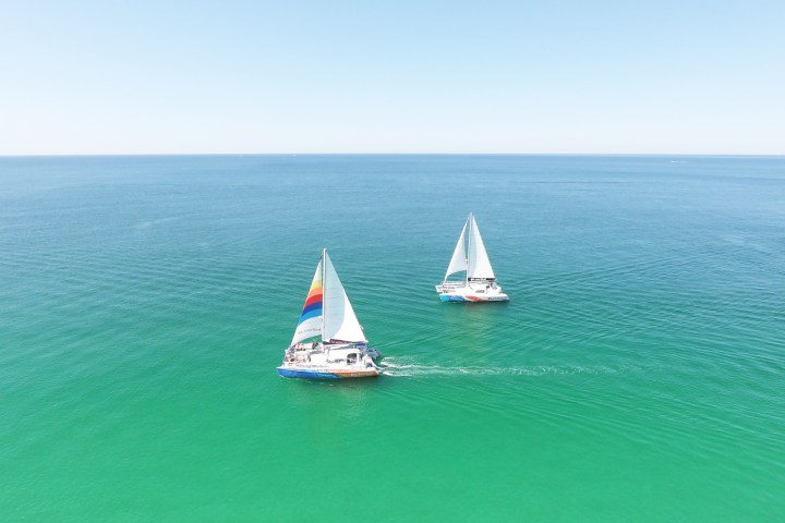 Two sailboats with colorful sails on calm, clear turquoise waters under a blue sky.