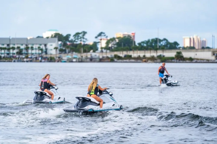 Three people riding jet skis on a calm body of water near a cityscape.