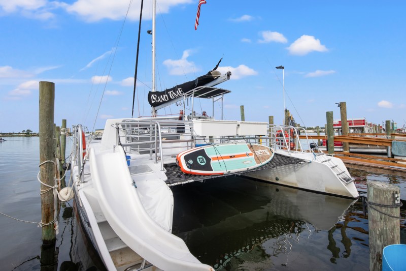 Docked white catamaran with paddleboard on deck, under a blue sky at a marina.