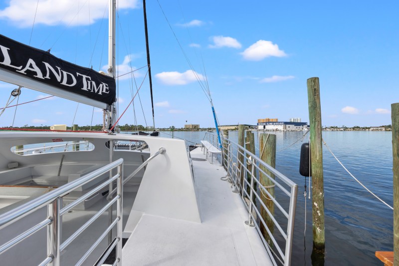 Docked sailboat with a black sail, metal railings, calm water, and buildings in the distance.