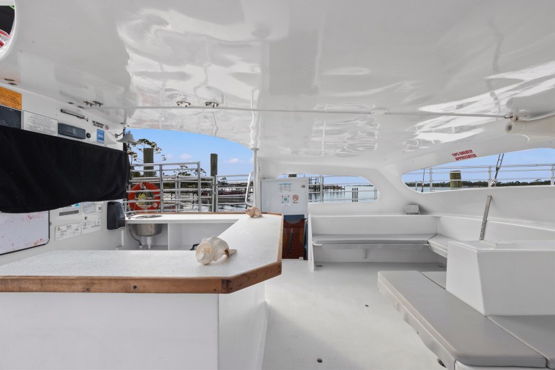 Interior of a boat with white counters and seating, dock visible through windows.
