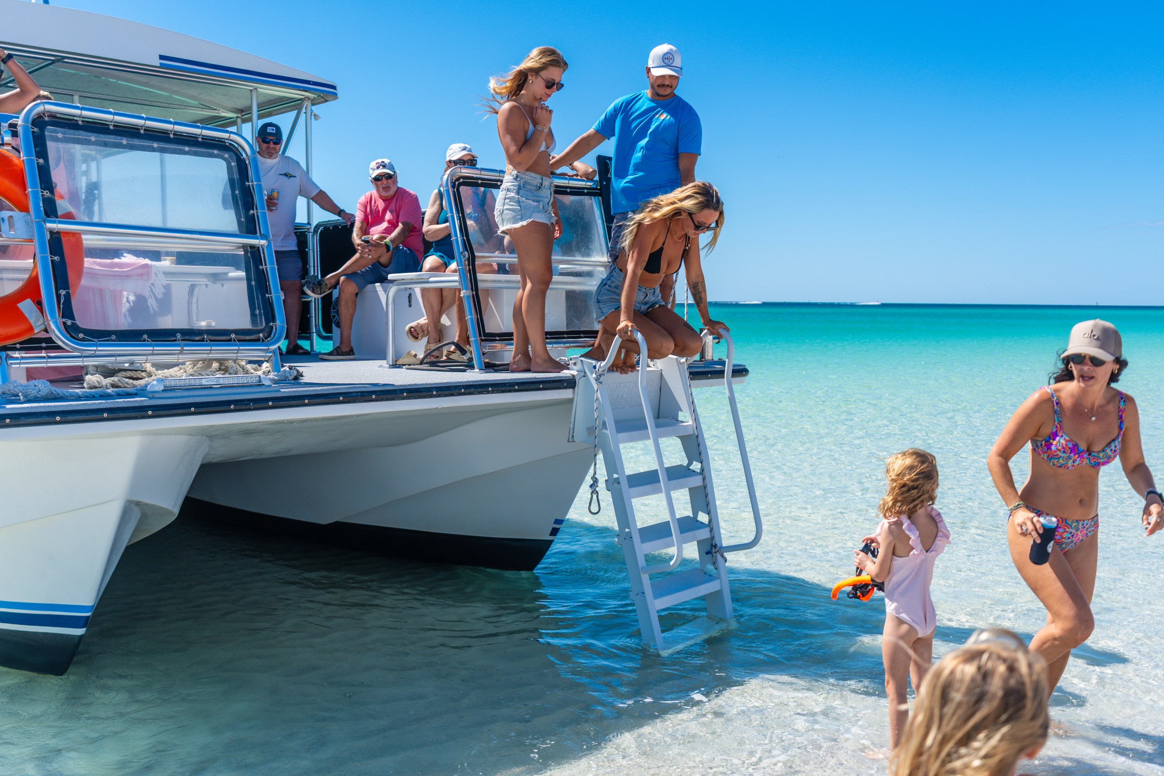 People disembark a boat onto a sandy beach with clear blue water under a sunny sky.