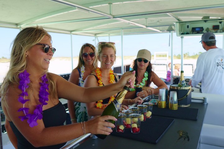 Four women in leis on a boat, one pouring drinks, with a bartender in the background.