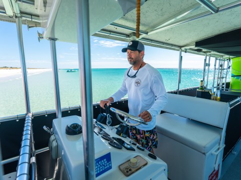 Man steering a boat in sunny coastal waters with beach in background.