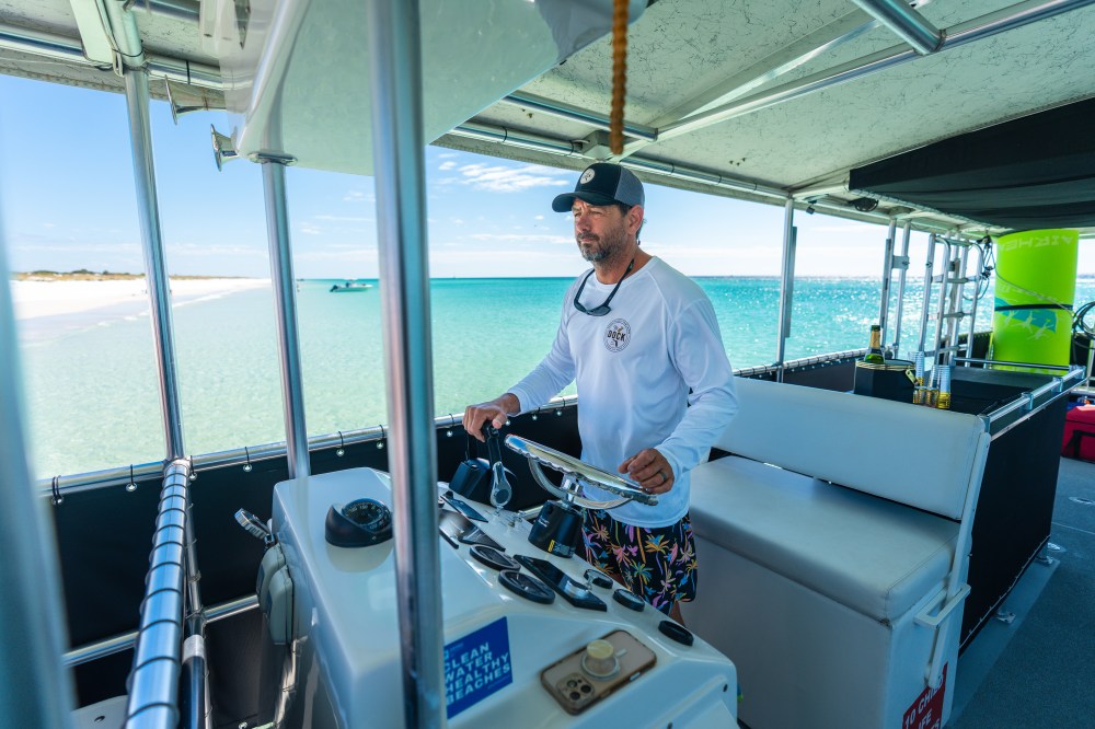 Man steering a boat in sunny coastal waters with beach in background.