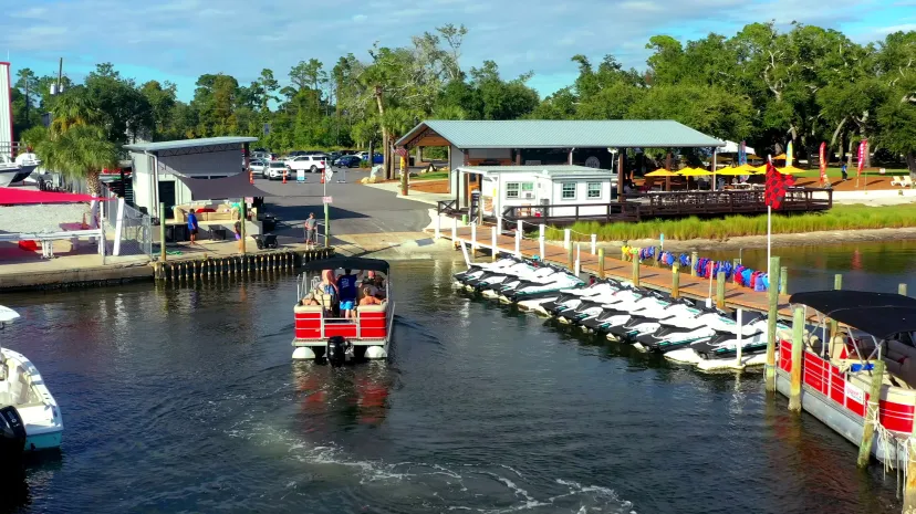 People on a red boat in a marina with docked jet skis and a waterfront building.
