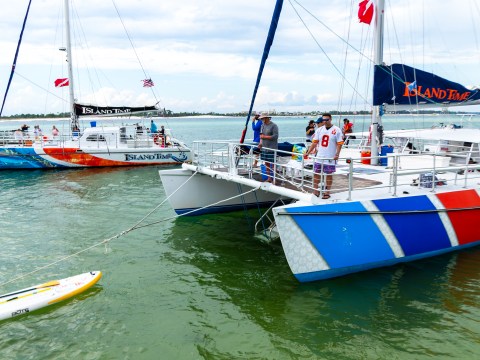 Colorful sailboats on water with people aboard and a paddleboard nearby.