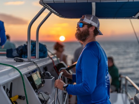Bearded man in blue shirt on a boat at sunset, steering the vessel.