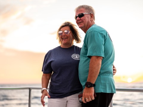 Two people smiling on a boat at sunset, wearing casual clothes and sunglasses.