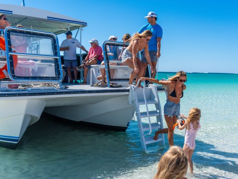 People disembarking a boat onto a sunny beach, with clear blue sky and ocean.