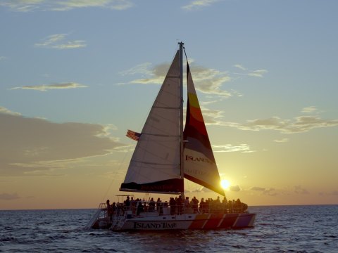 Sailboat on ocean at sunset with silhouetted people on board.