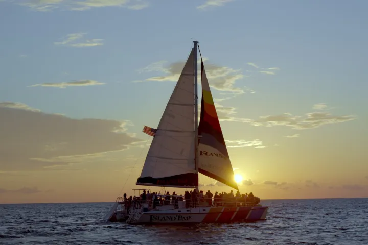 Sailboat on ocean at sunset with silhouetted people on board.
