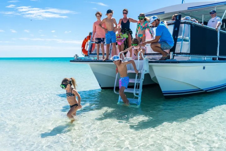 People on a boat, with children stepping into clear turquoise water on a sunny day.