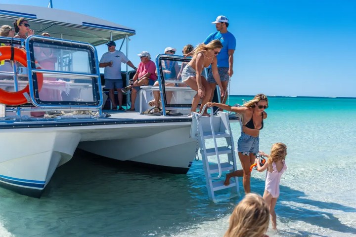 People disembarking a boat onto a sunny beach, clear skies above.