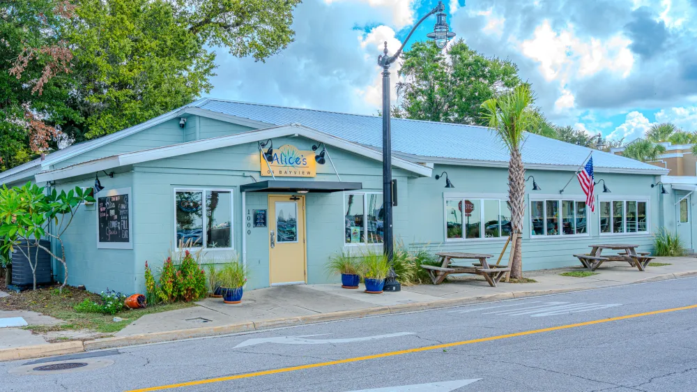 Light blue diner with outdoor seating, American flag, and greenery on a cloudy day.