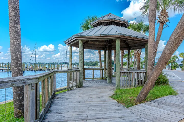 Wooden gazebo on a lakeside boardwalk with palm trees and blue sky.